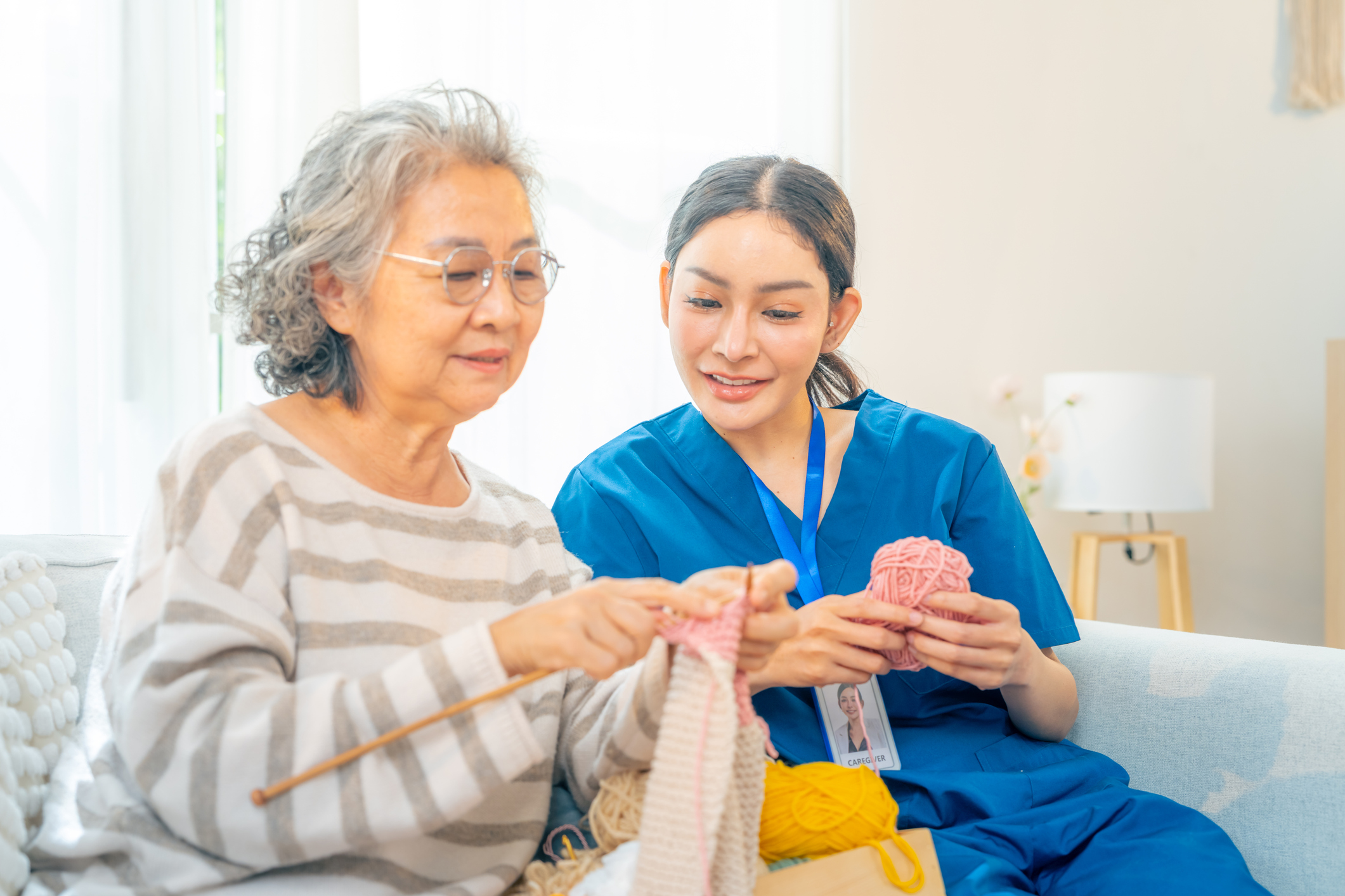 Alzheimer’s caregiver knits alongside an older woman on a living room couch.