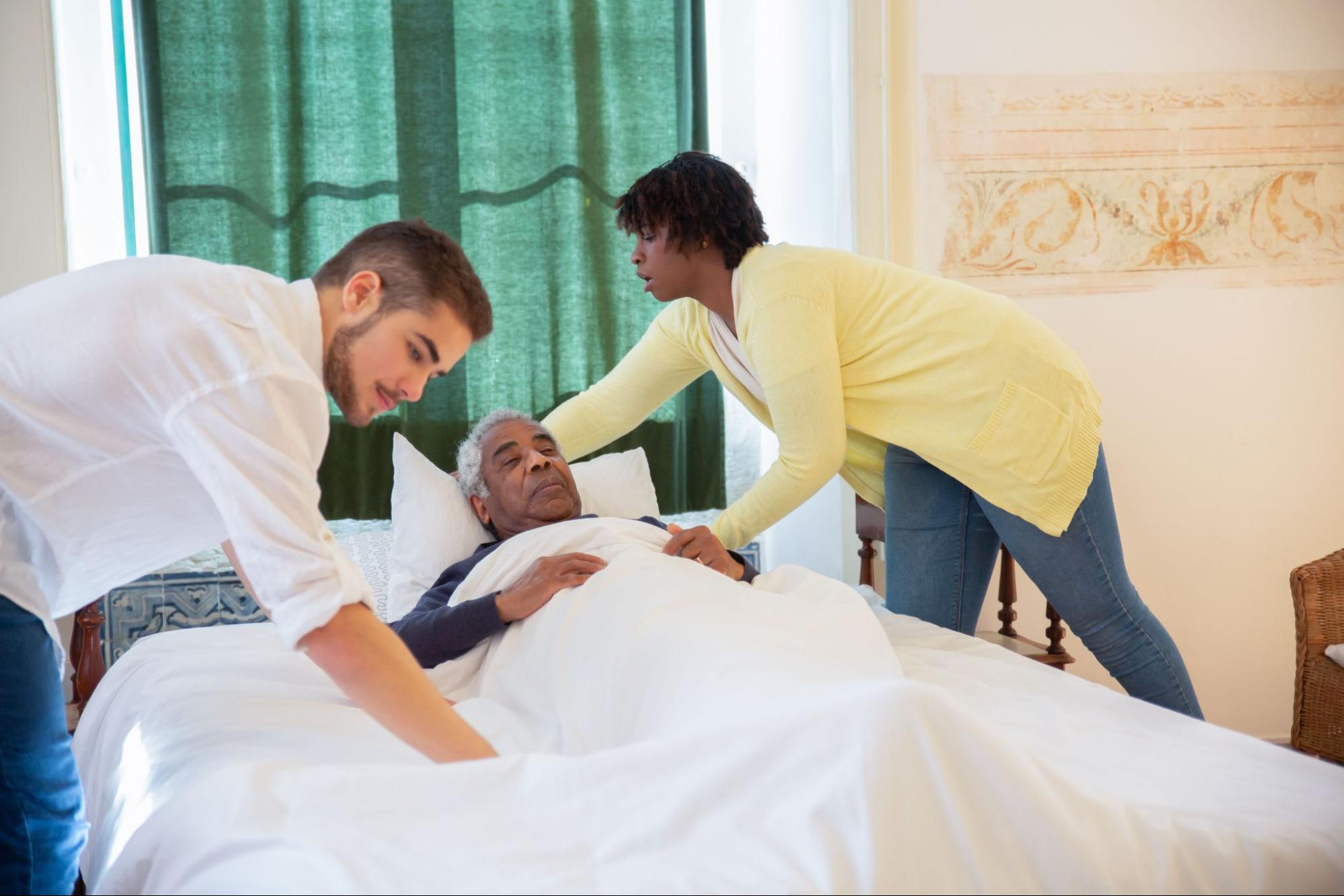 A man and a woman tuck an older man into bed for overnight home care.